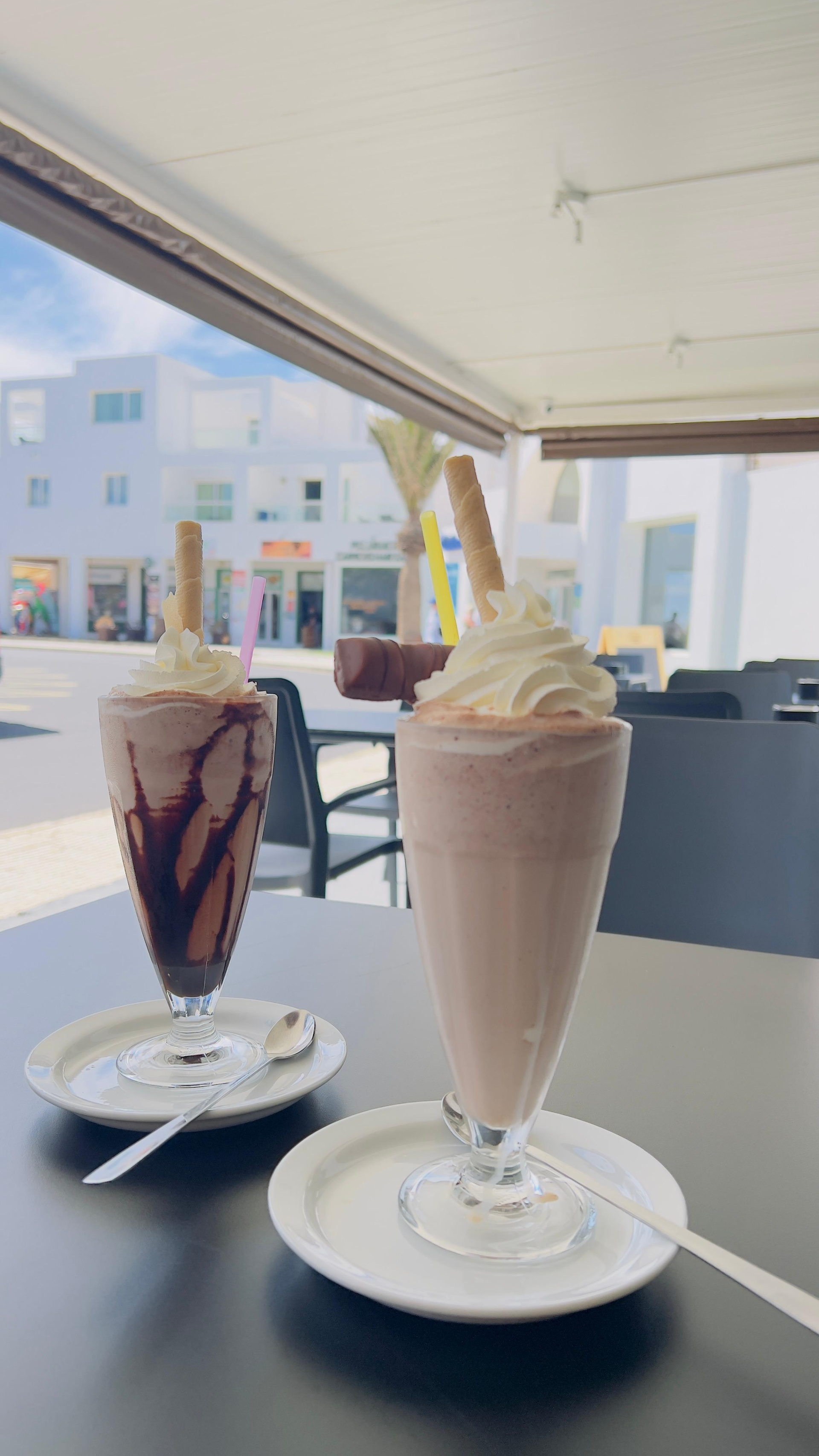 Dos bebidas heladas en platillos en una cafetería al aire libre, una de chocolate y otra con crema batida.