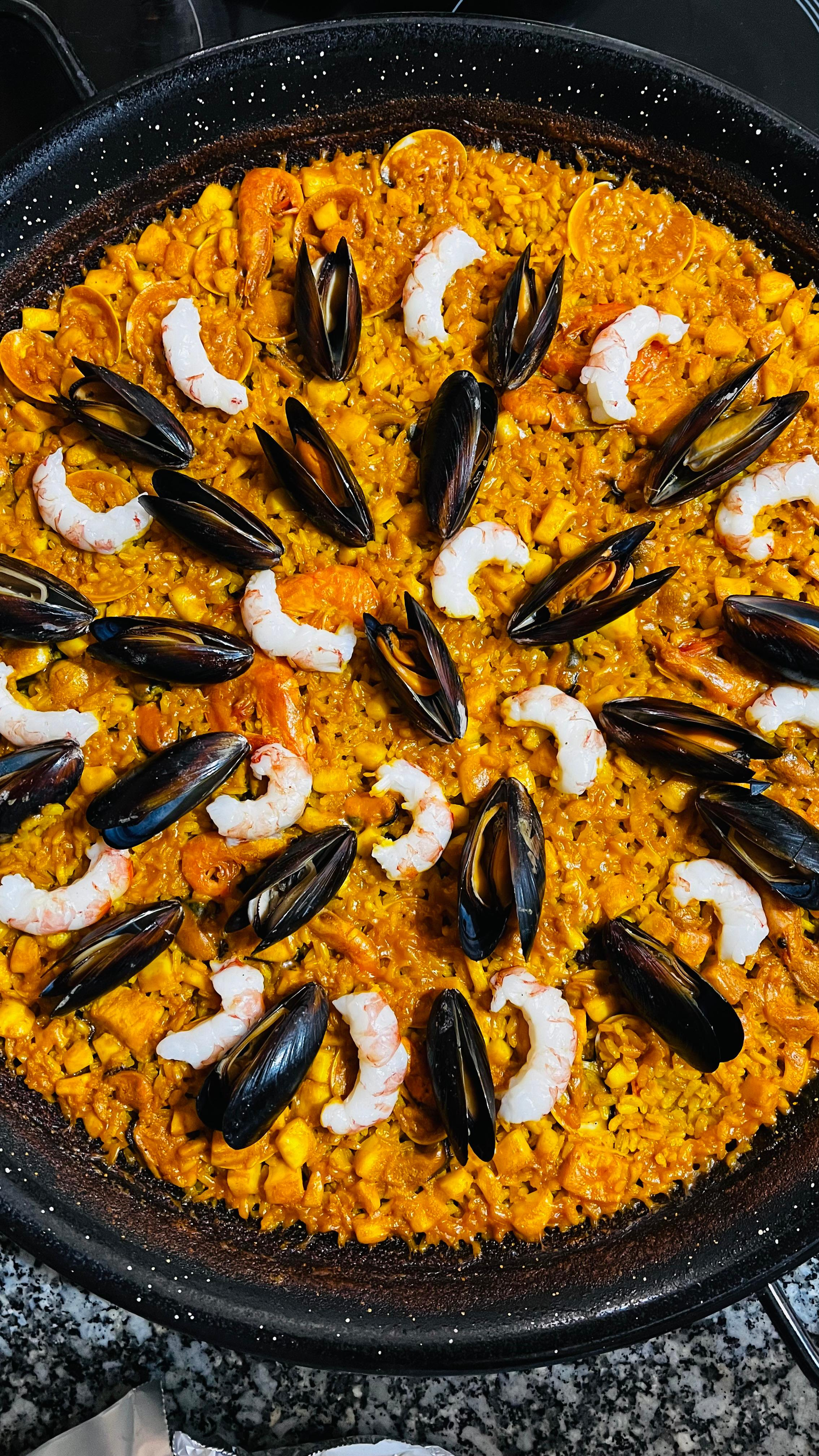 Paella en una sartén negra, cubierta con mejillones y gambas, acompañada de arroz naranja y rodajas de limón.