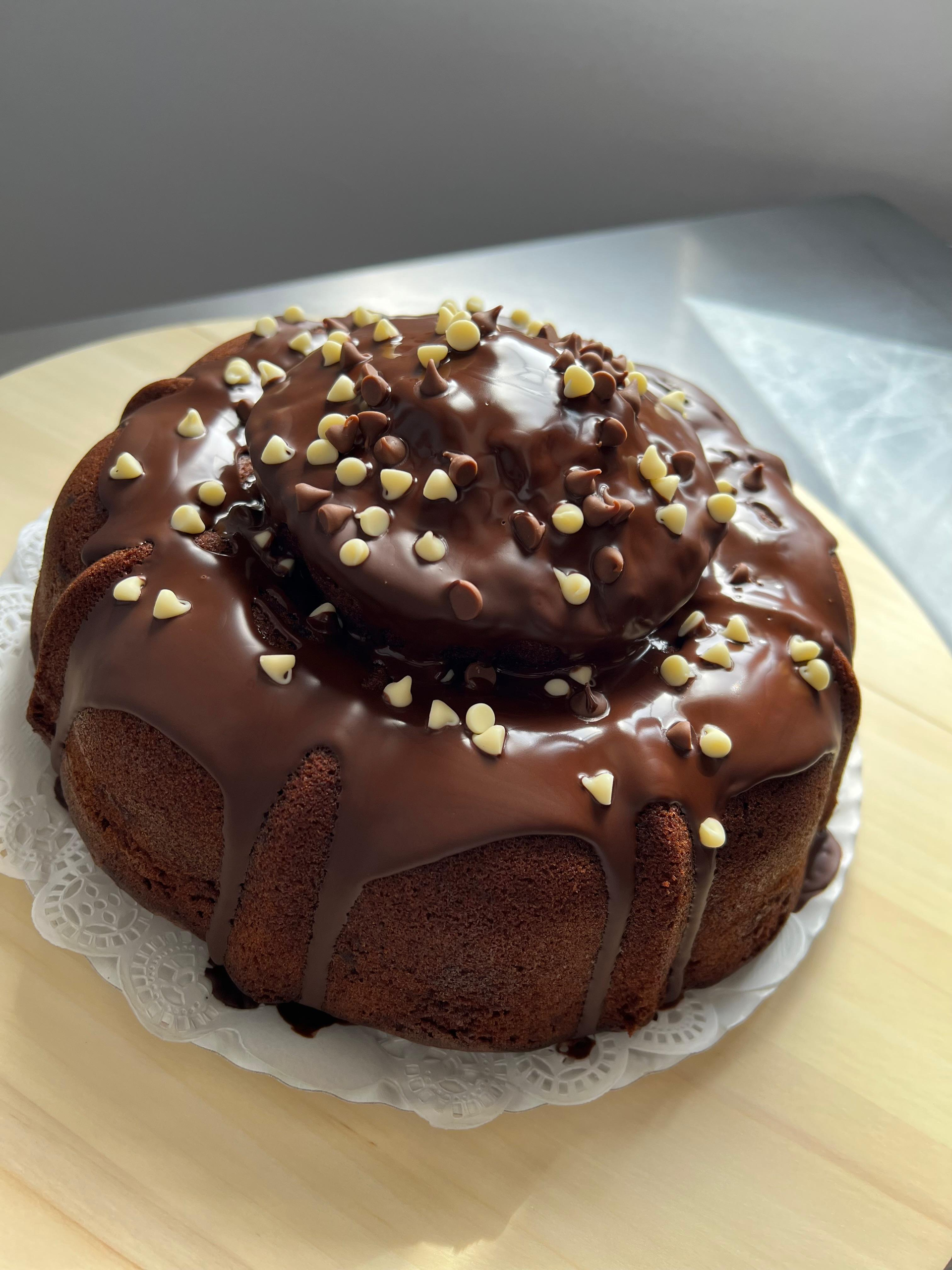 Pastel Bundt glaseado con chocolate y nueces picadas sobre un tapete blanco, sobre una mesa de madera clara.