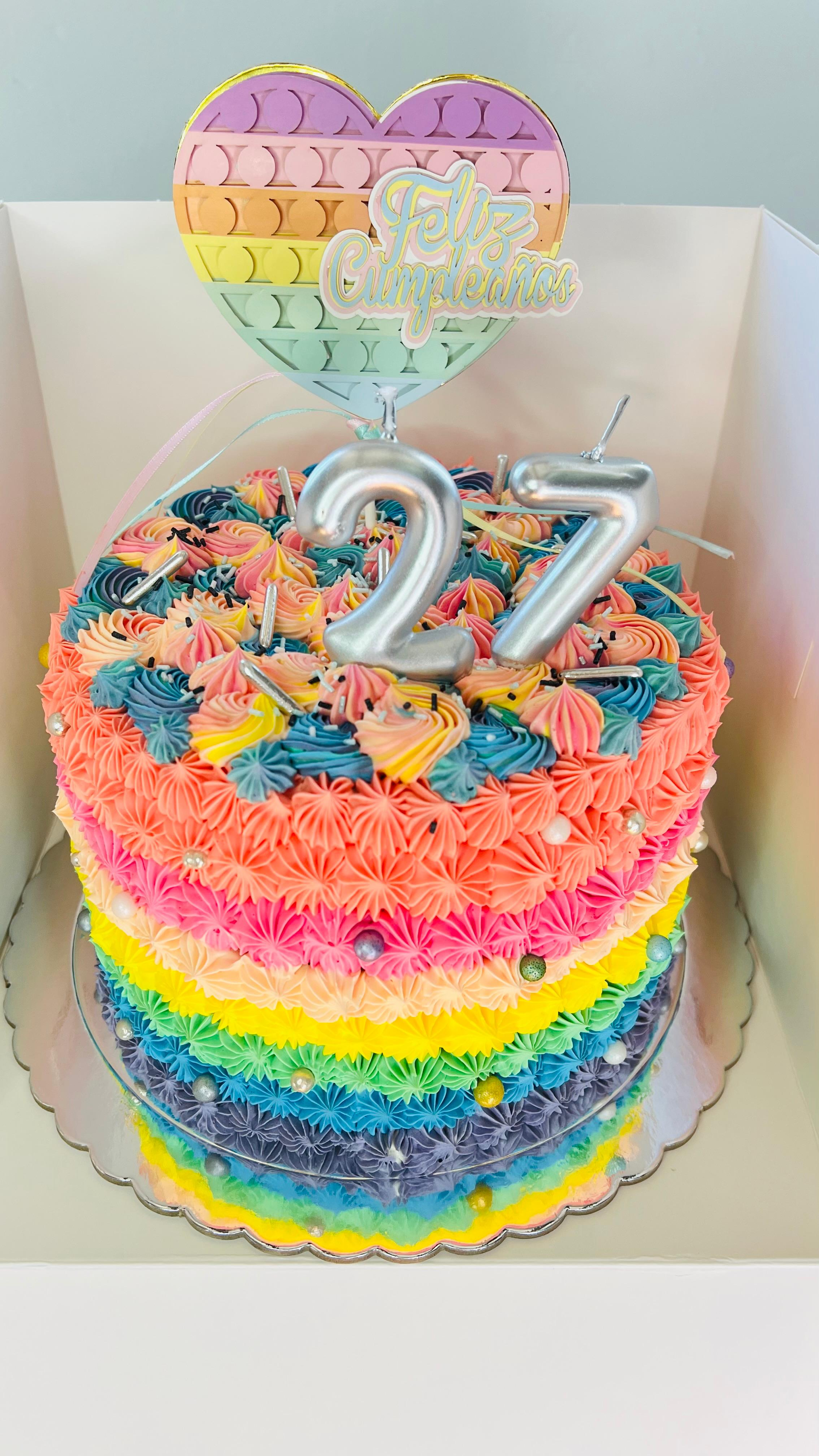 Tarta arcoíris colorida con adorno de globo en forma de corazón y velas plateadas con el número 27.