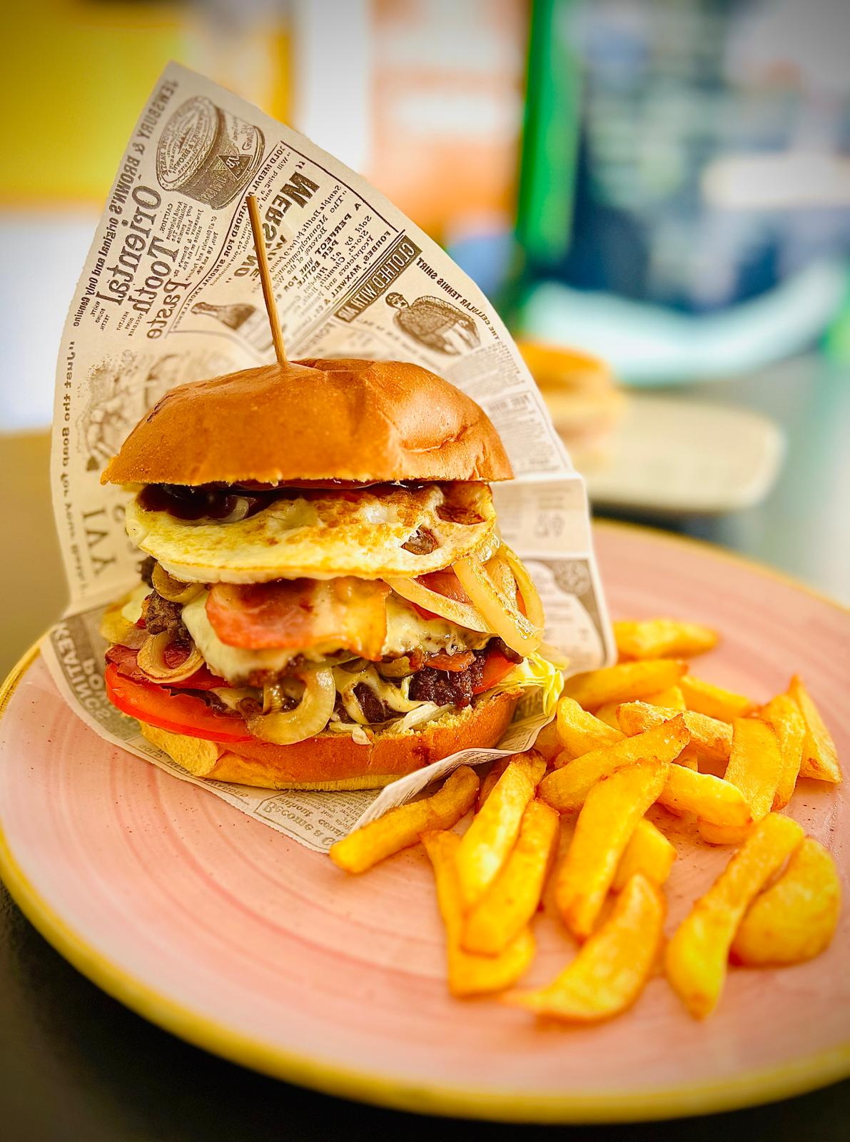 Hamburguesa y patatas fritas en un plato rosa con envoltorio estilo periódico en un luminoso ambiente de restaurante.