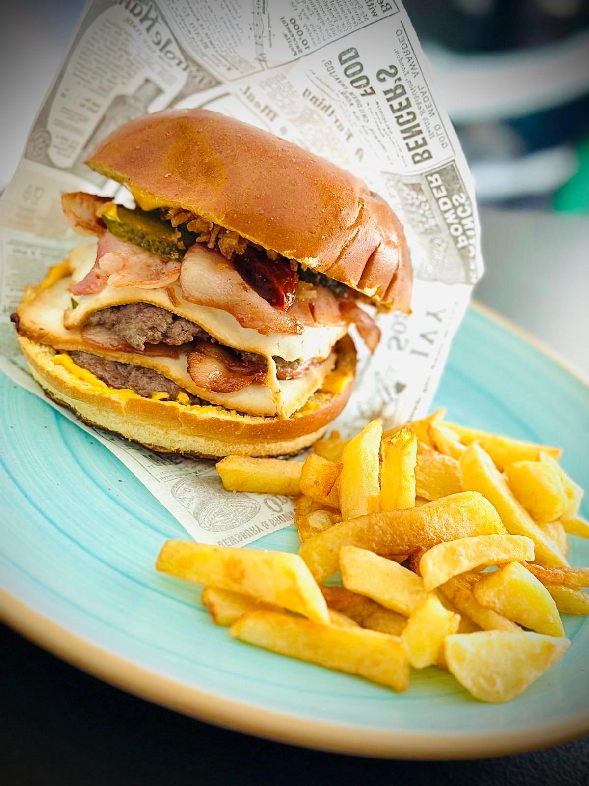 Hamburguesa y patatas fritas en un plato, envuelto en papel estilo periódico, con un fondo turquesa.