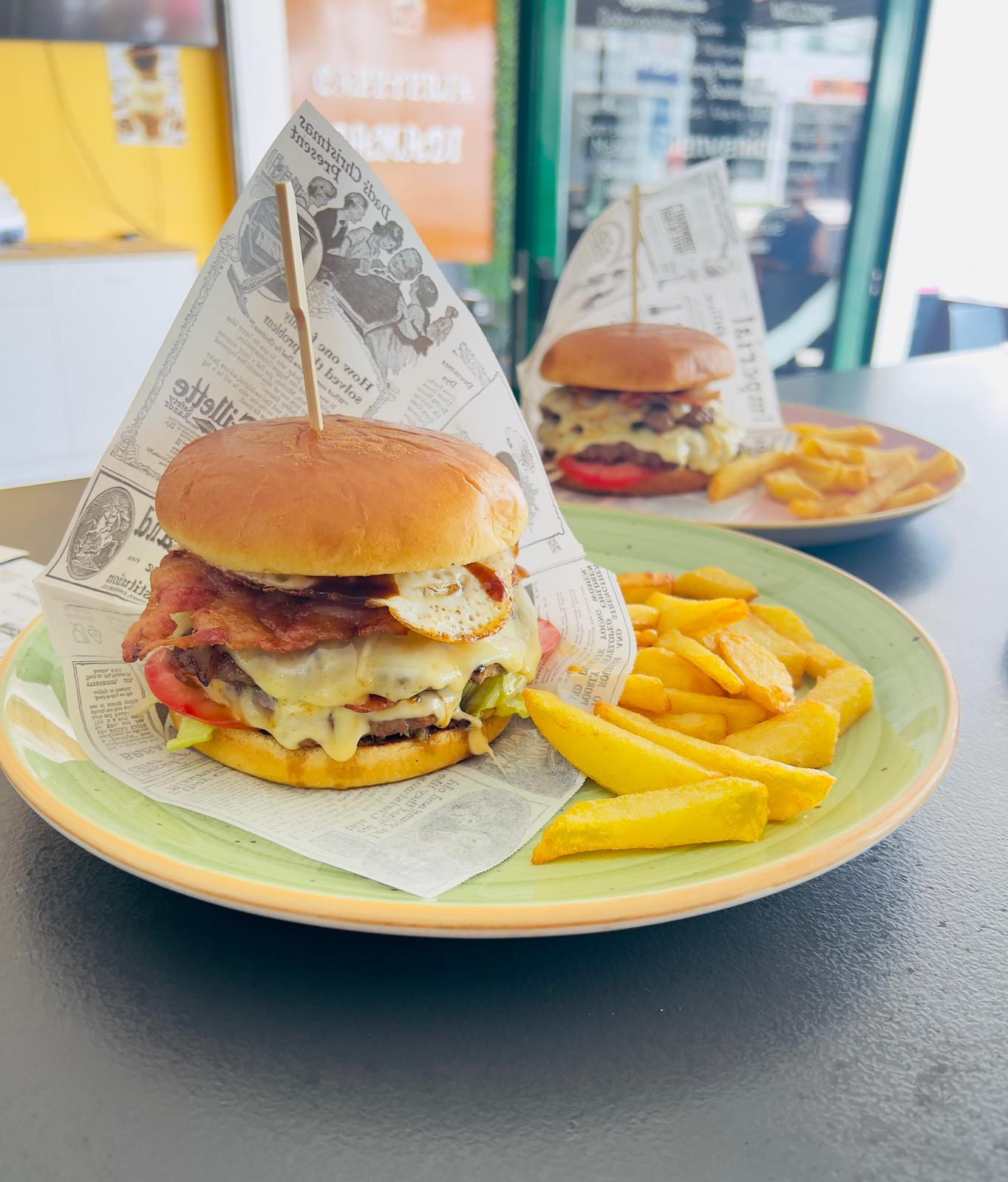 Dos hamburguesas y papas fritas en platos verdes sobre una mesa de café, con los envoltorios de las hamburguesas.