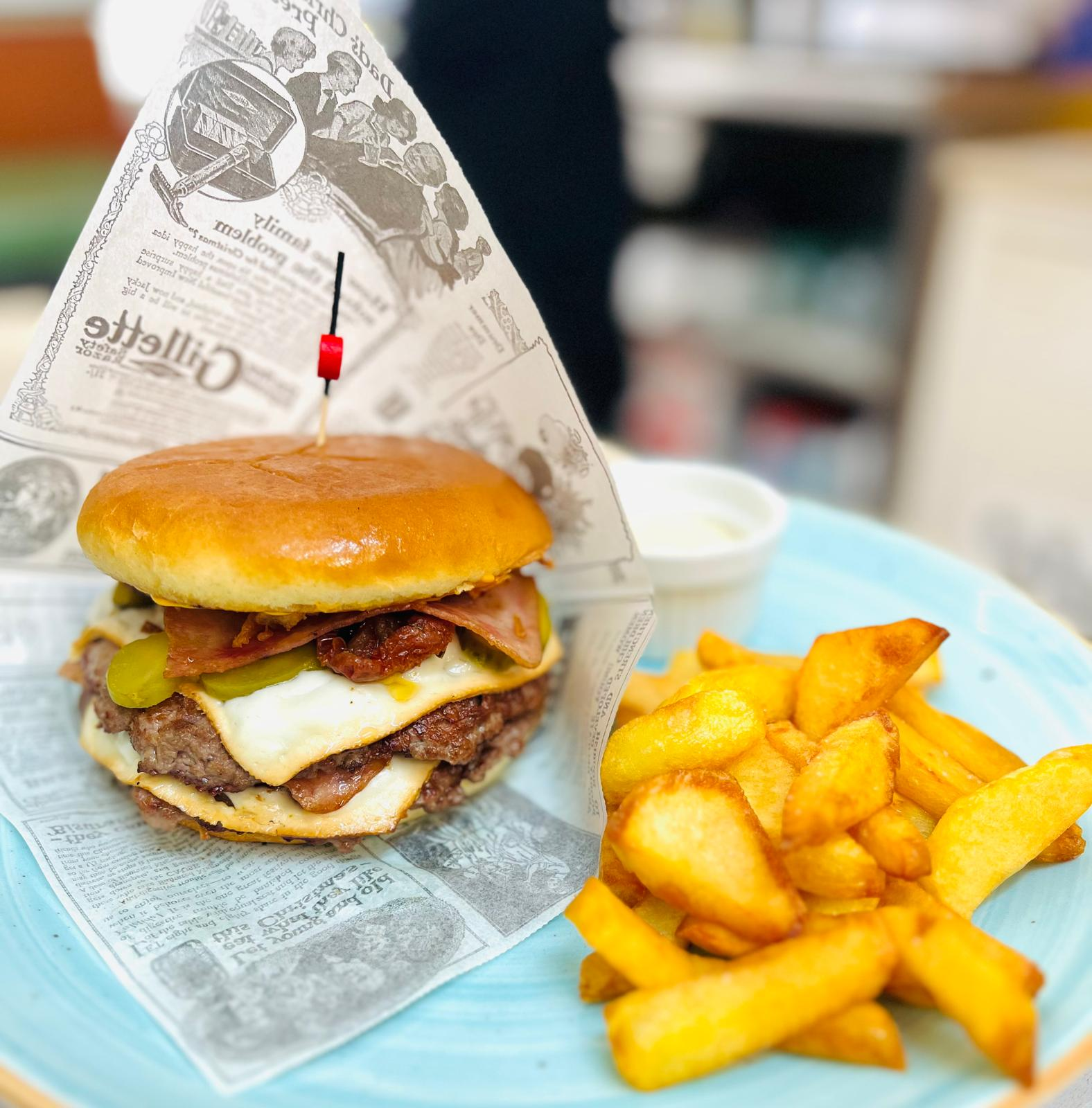 Hamburguesa y patatas fritas en una bandeja azul con forro de papel y un pequeño vaso de bebida al fondo.