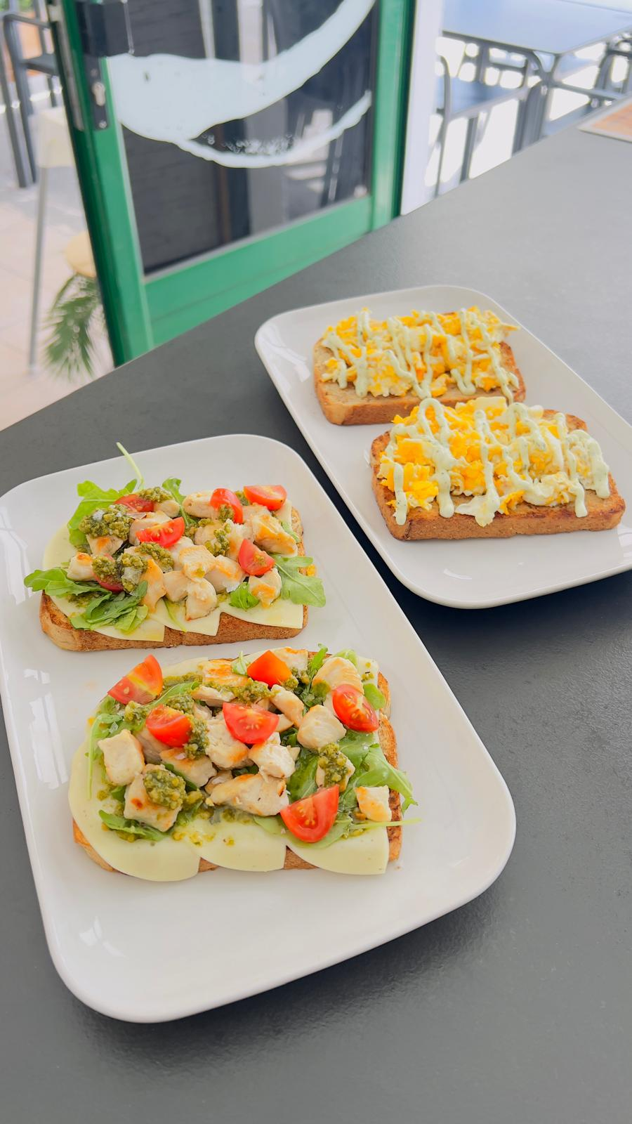Tres sándwiches abiertos con verduras, servidos en un plato sobre una mesa gris junto a una ventana.