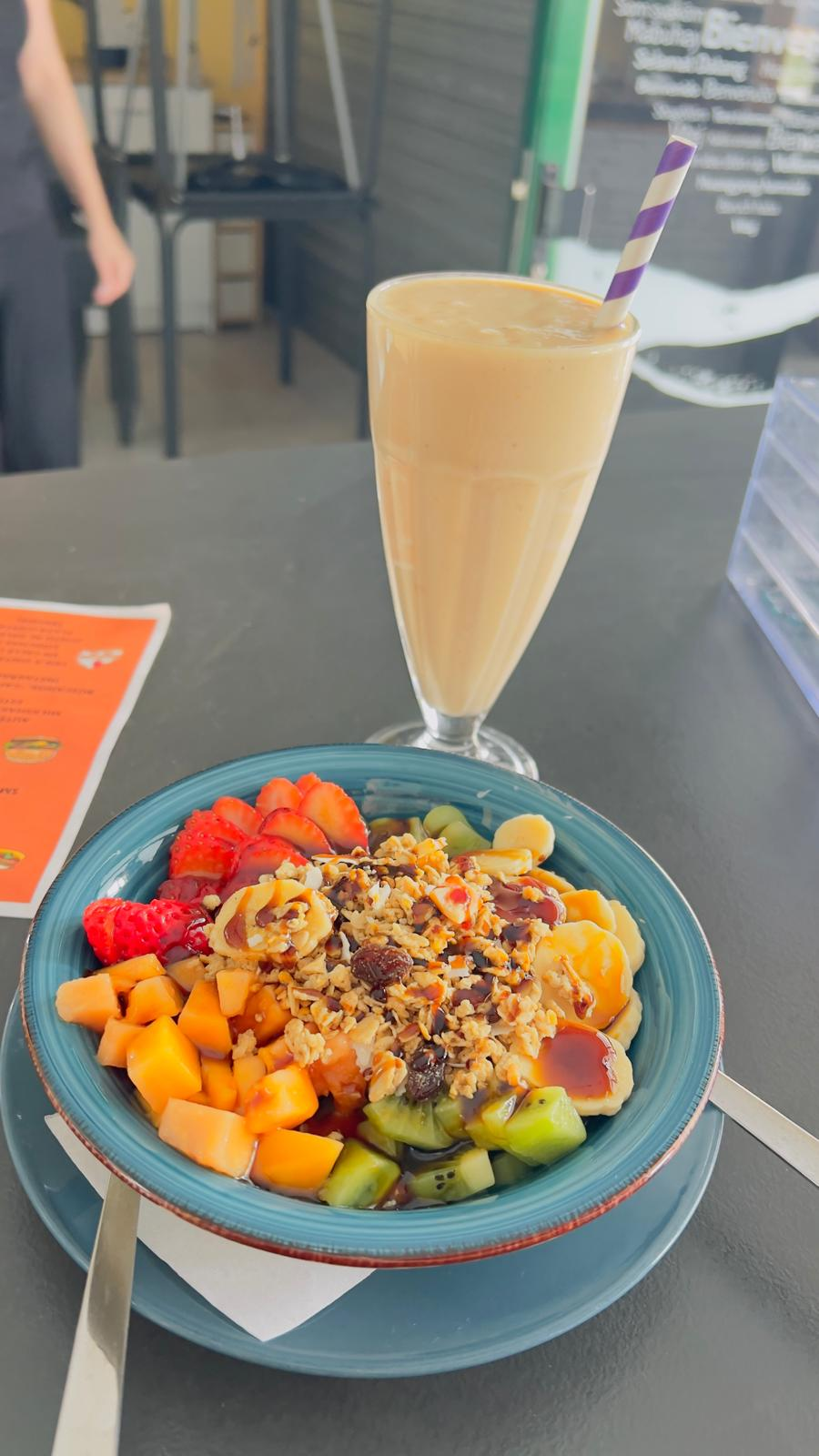 Batido y tazón de frutas con kiwi, fresas, mango y granola en una mesa de cafetería.