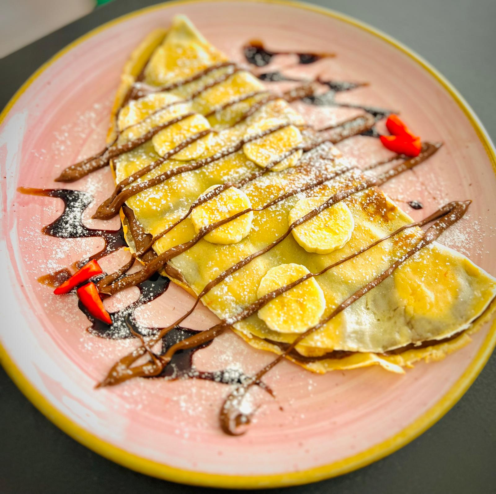Crepe doblada bañada en chocolate sobre un plato rosa, espolvoreada con azúcar glas y acompañada de fresas.