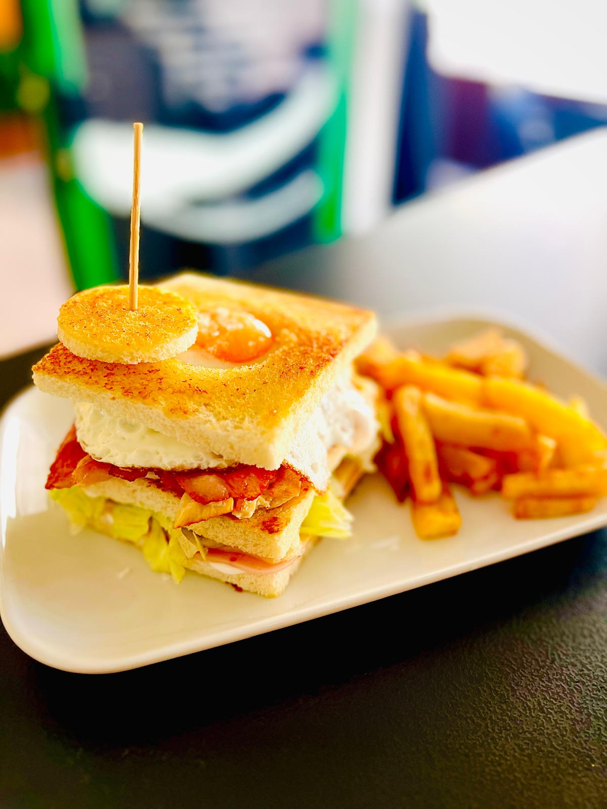 Sándwich club y patatas fritas en un plato blanco, coronado con un pincho, sobre una mesa oscura.