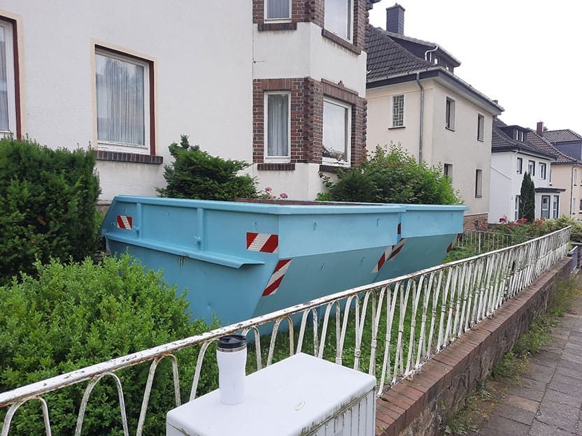 Vor einem weißen Haus steht ein blauer Müllcontainer.