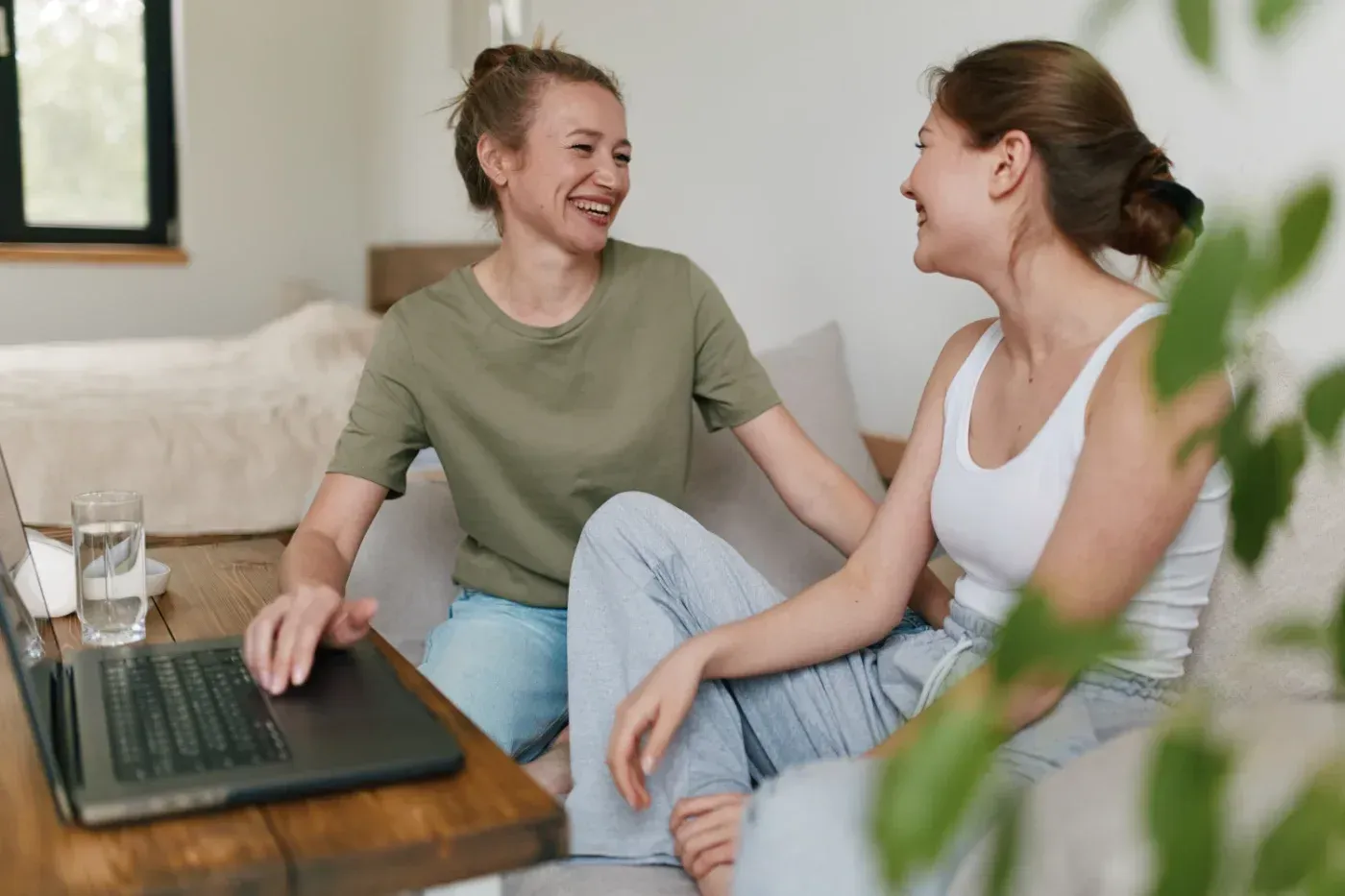 Zwei Frauen lachen auf einem Sofa, eine von ihnen benutzt einen Laptop. Natürliches Licht durchflutet den Raum.