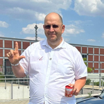 Manfred Kremer in einem weißen Hemd hält ein Einmachglas, zeigt nach oben und steht vor einem Gebäude, im Hintergrund ist ein hoher Turm zu sehen.