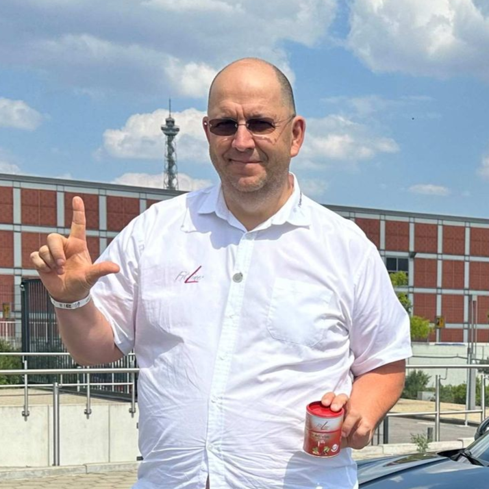 Manfred Kremer in einem weißen Hemd hält ein Einmachglas, zeigt nach oben und steht vor einem Gebäude, im Hintergrund ist ein hoher Turm zu sehen.