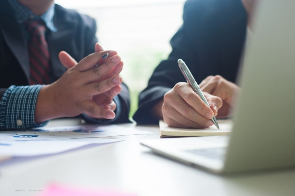 Zwei ineinander verschränkte Hände mit Kugelschreiber zwischen Zeige- und Mittelfinger, rechts davon Hand, die mit Stift ein Schriftstück unterzeichnet