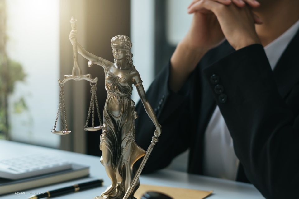 Metall-Skulptur Justitia, dahinter auf Tisch abgestützte Ellbogen mit in der Luft ineinander verschränkten Händen eines Mannes in Anzug 