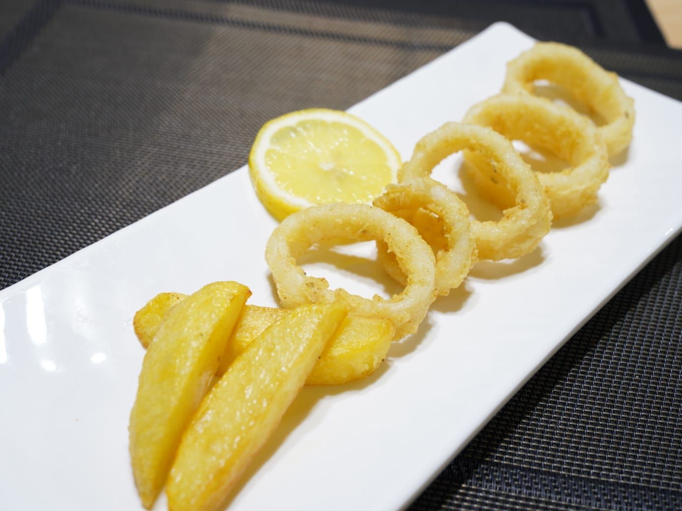 Anillos de calamares fritos y gajos de papa en un plato blanco con una rodaja de limón, sobre una superficie oscura.