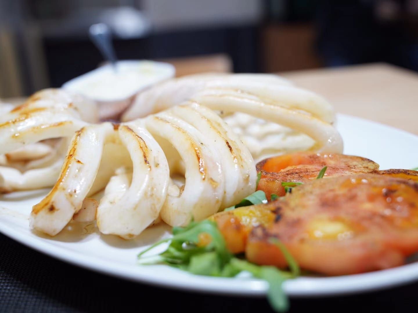 Anillos de calamar y tomates a la parrilla en un plato blanco, adornados con verduras y una guarnición de salsa.