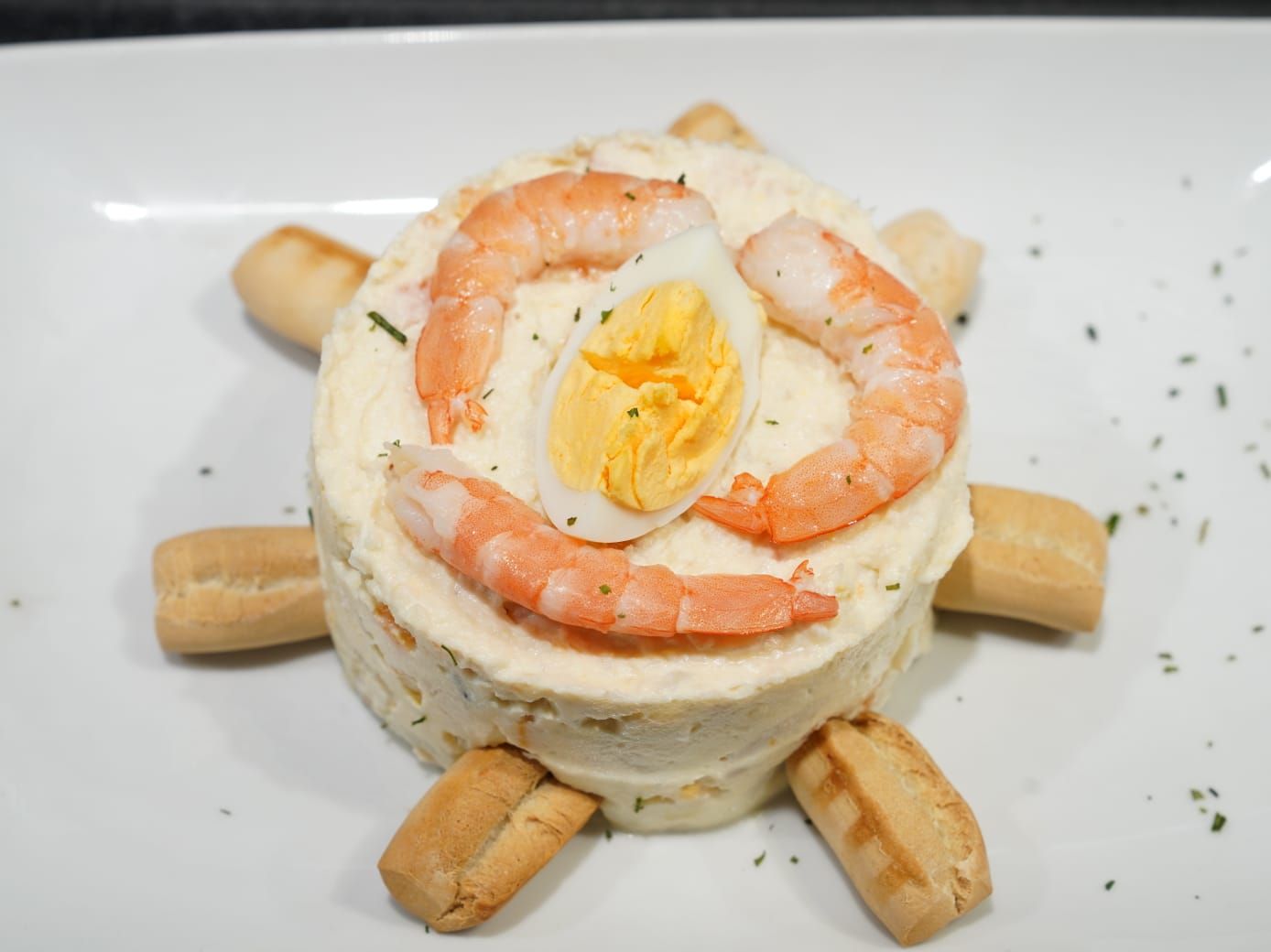 Una ensalada cremosa con forma de tortuga, decorada con camarones, un huevo cocido y palitos de pan en un plato blanco.