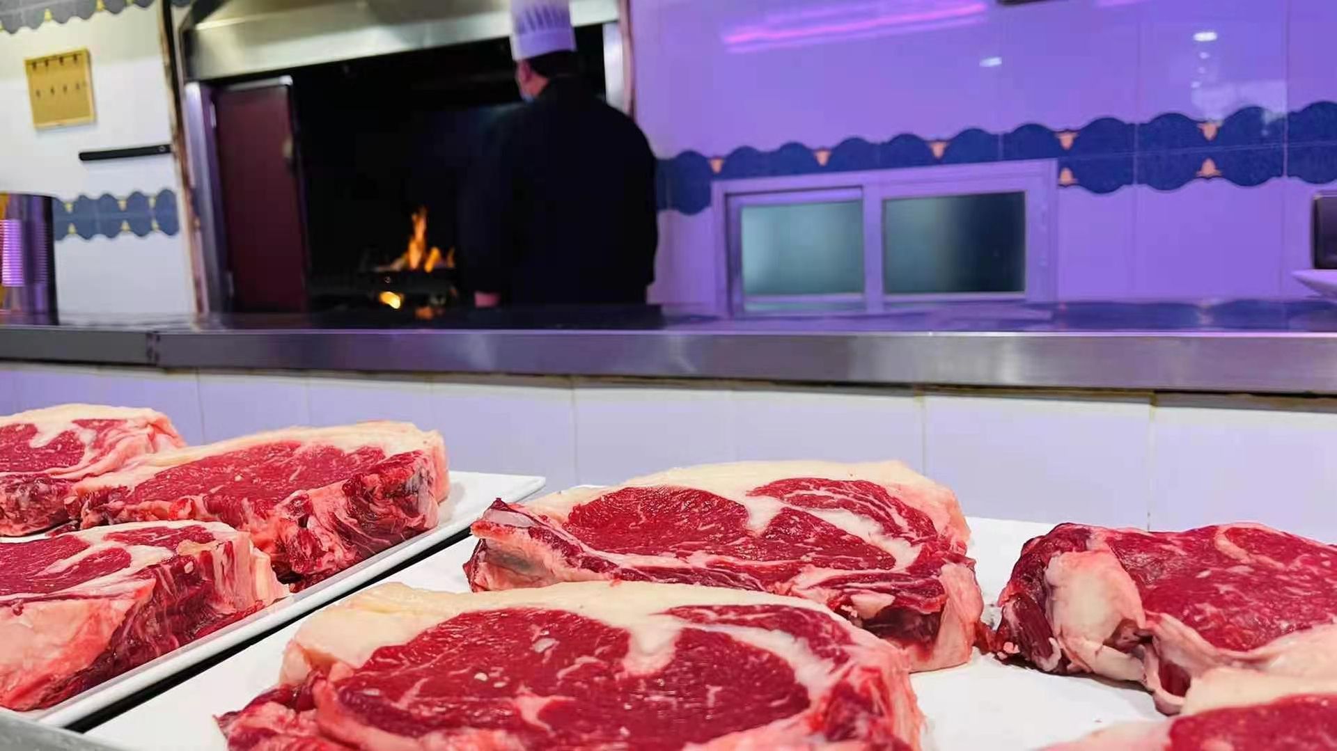Filetes crudos en exhibición en el mostrador de un restaurante, con un chef vigilando el fuego en el fondo.