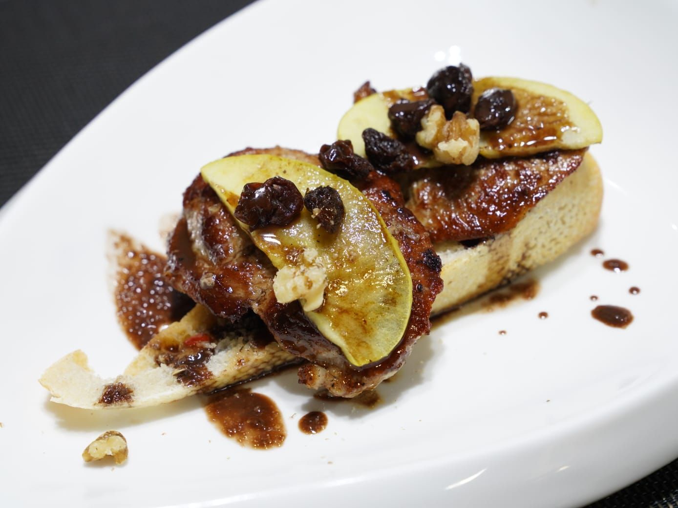 Tostada de foie gras cubierta con rodajas de manzana, nueces y pasas, rociada con salsa en un plato blanco.