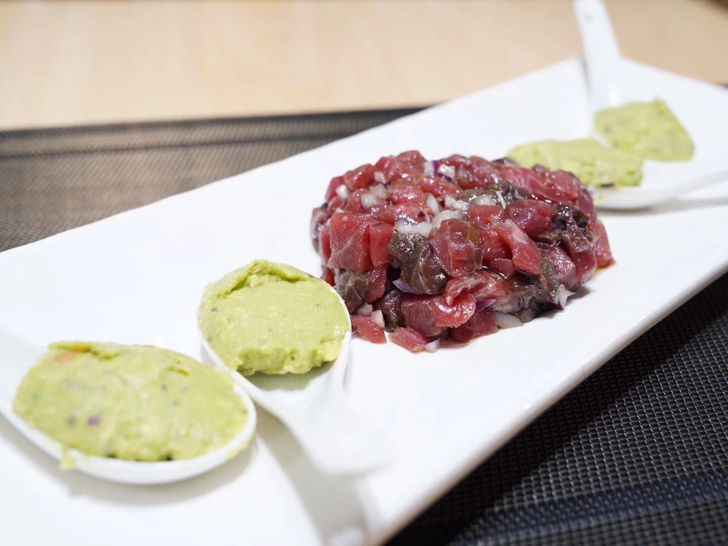 Tártaro de ternera con dos cucharadas de puré de aguacate verde servido en un plato rectangular blanco.