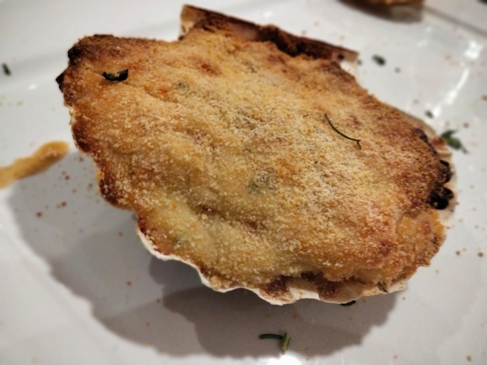 Vieira horneada en su concha en un plato blanco, cubierta con pan rallado y hierbas.