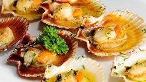 Vieiras cocidas en sus conchas, adornadas con hierbas, en un plato blanco.