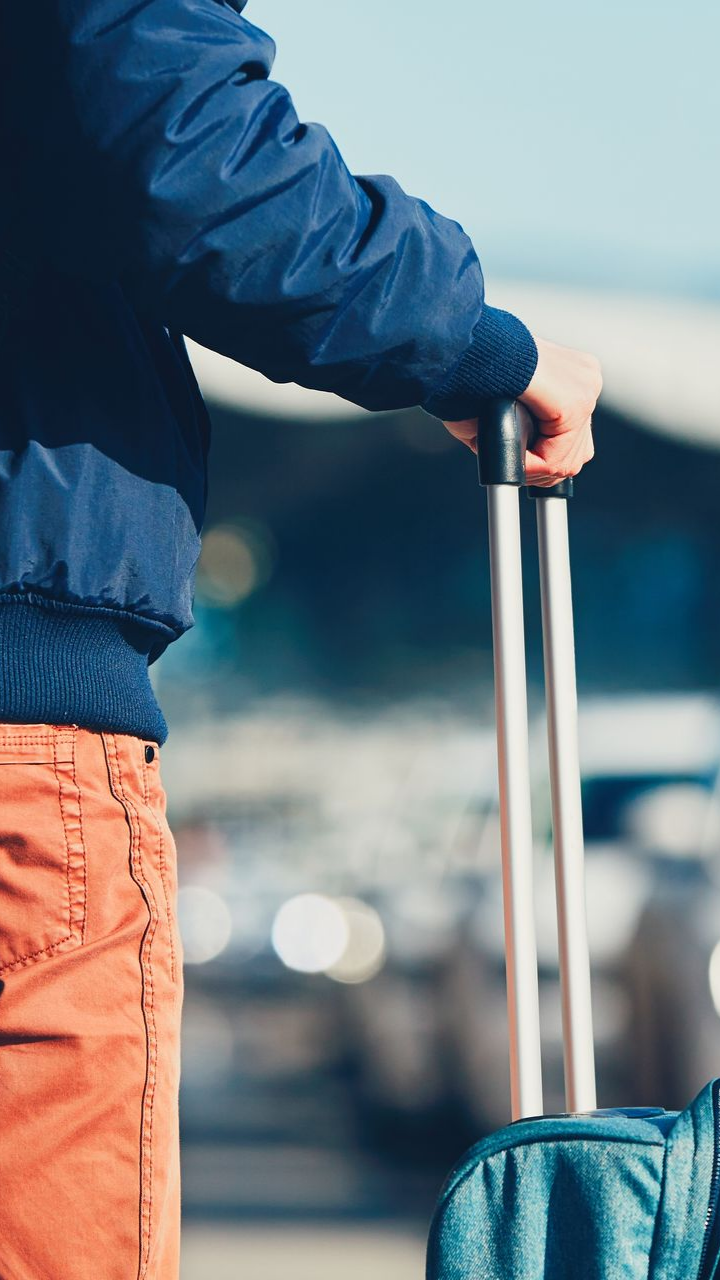 Une personne vêtue d'une veste bleue et d'un pantalon orange se tient devant un terminal d'aéroport, tenant la poignée d'une valise à roulettes.