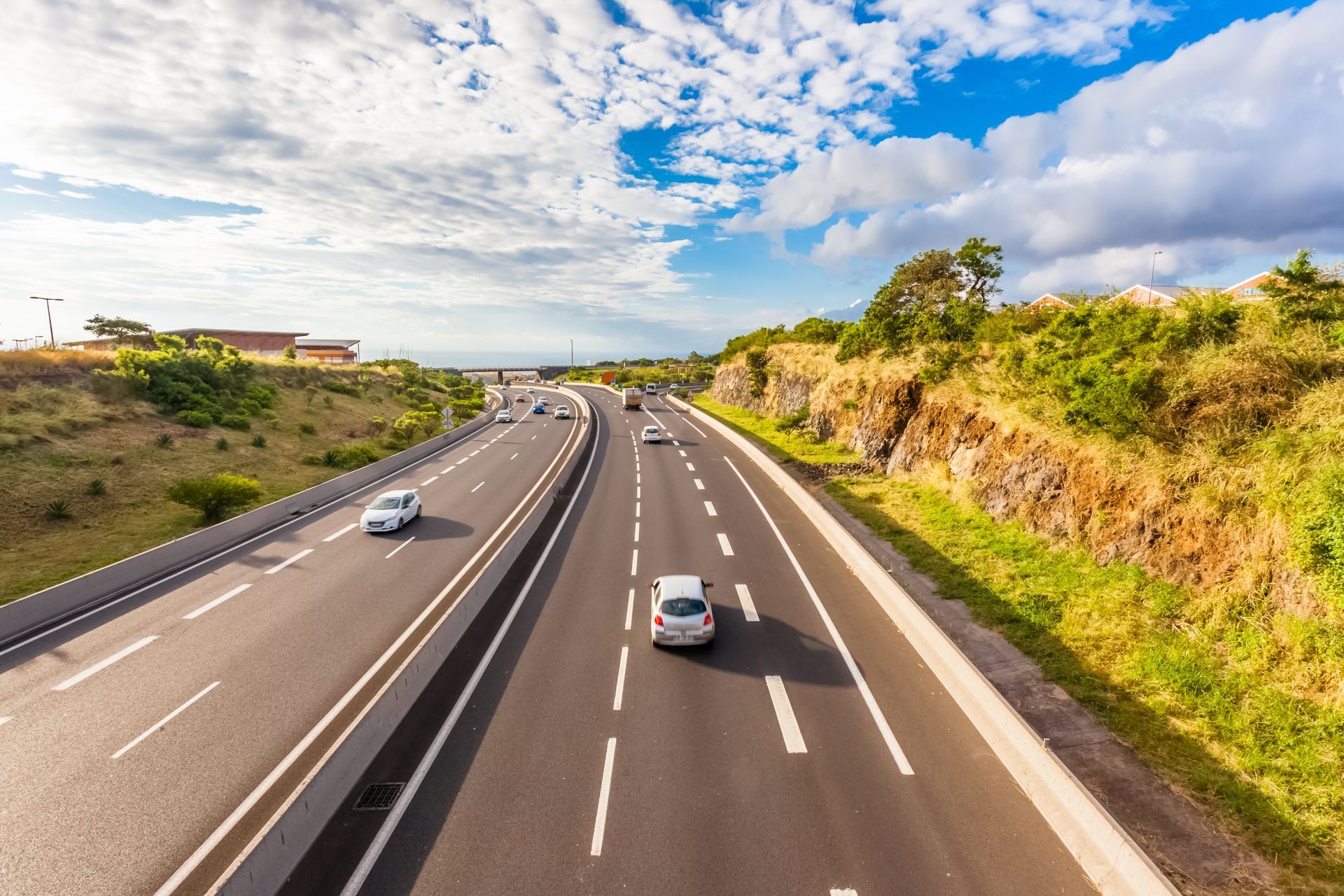 Vue ensoleillée en plongée d'une autoroute à plusieurs voies serpentant à travers un paysage verdoyant et rocailleux, avec une circulation automobile légère.
