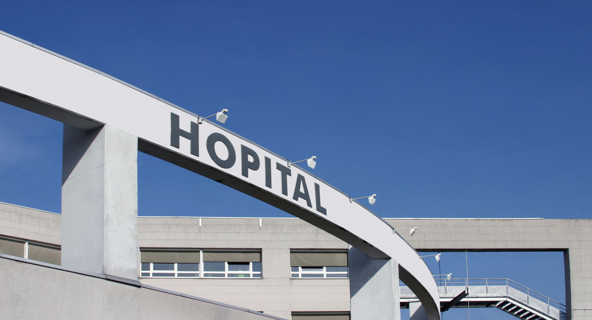 Une arche blanche avec le mot HÔPITAL en lettres noires sur un ciel bleu clair, masquant partiellement un bâtiment hospitalier.