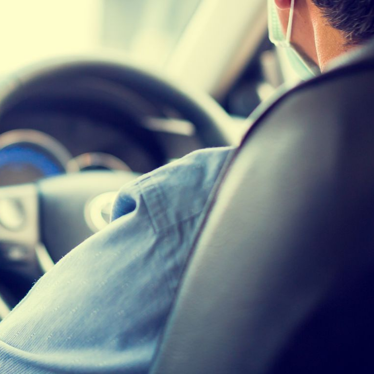 Une personne portant un masque conduit une voiture, vue depuis l'arrière du siège conducteur.