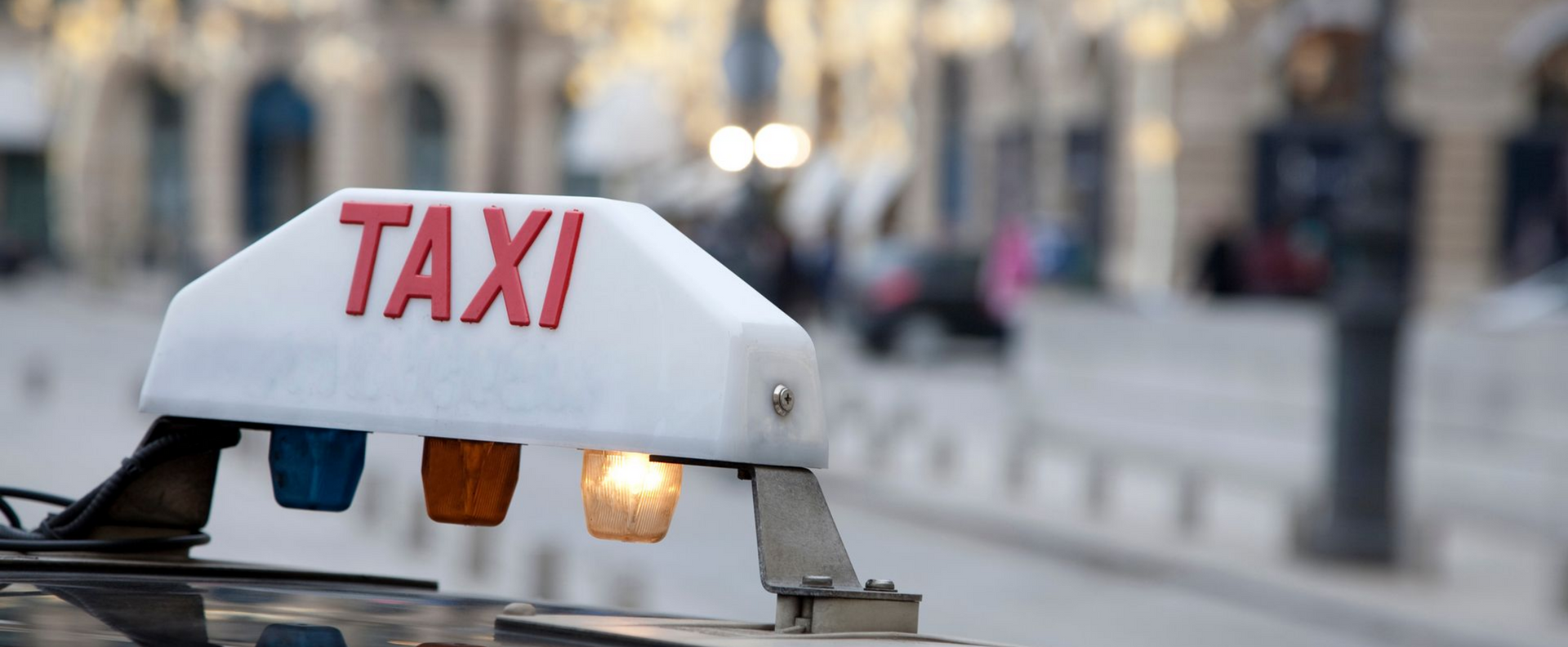 Gros plan sur un panneau de toit de taxi avec le mot TAXI en rouge, comportant trois petites lumières en dessous, dont une est allumée.
