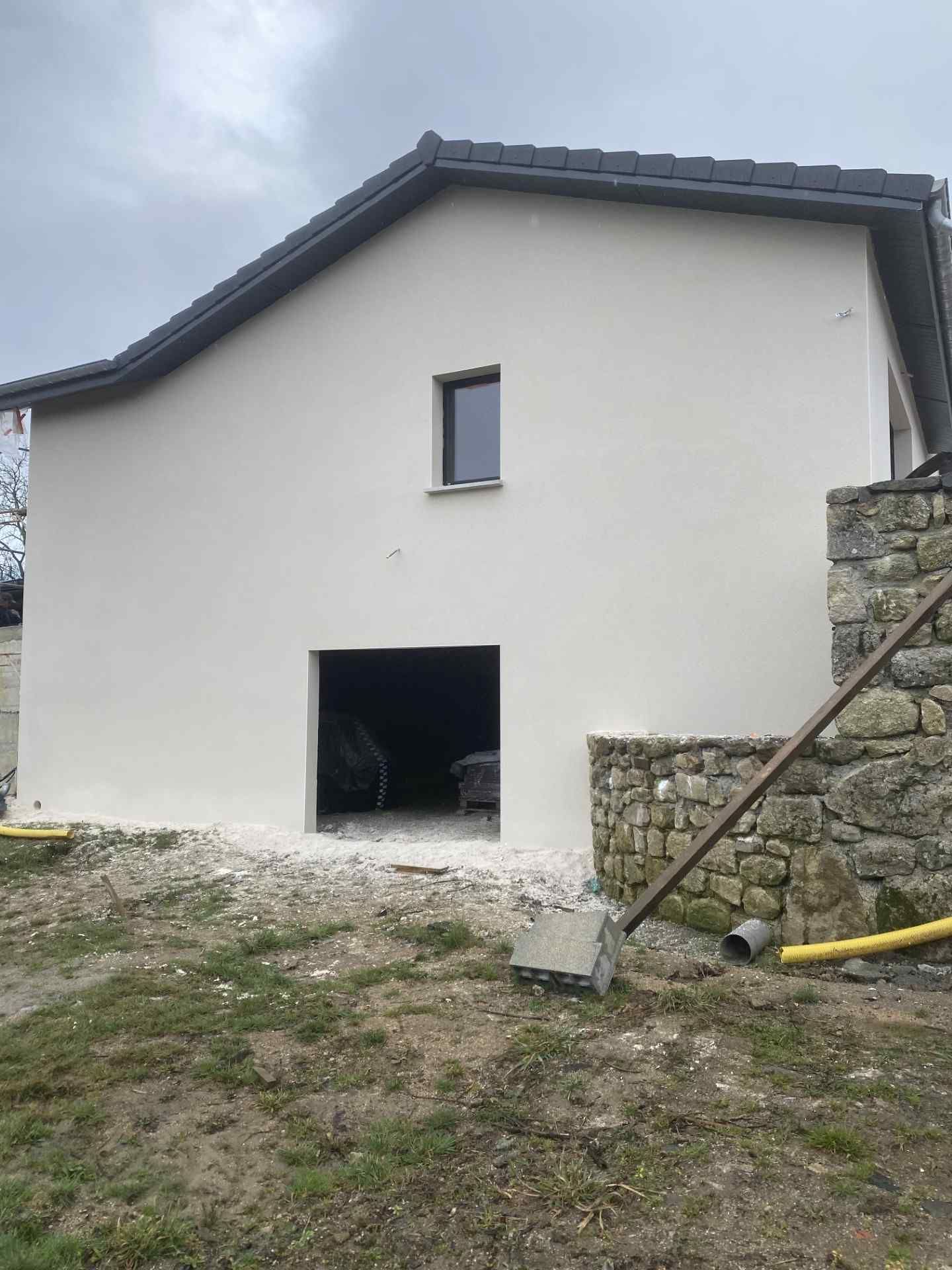 Extérieur d'une maison blanche avec une porte ouverte de type garage, un mur de soutènement en pierre et une cour en gravier sous un ciel couvert.