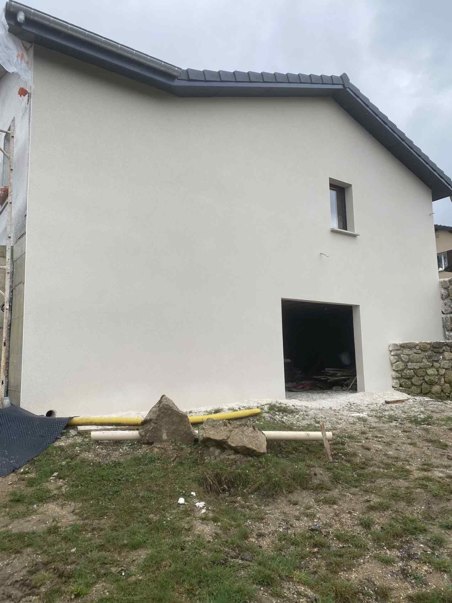 Extérieur d'une maison blanche avec un toit gris, une petite fenêtre et une porte ouverte, vus depuis une cour gazonnée.