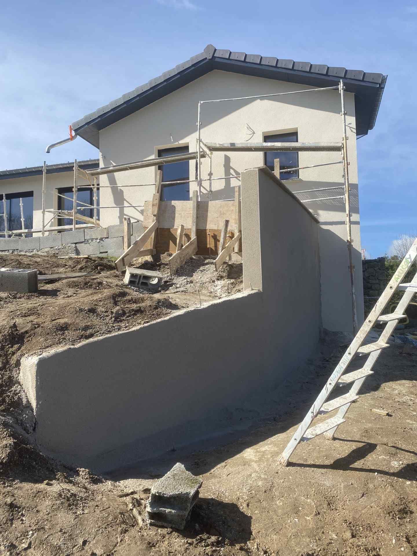 Maison en construction avec mur en béton inachevé et échelle sur un terrain en terre battue sous un ciel bleu