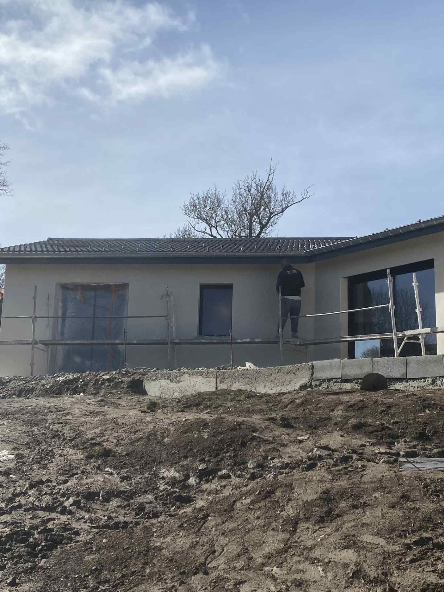 Maison de plain-pied en construction, avec échafaudages et terrain boueux devant.
