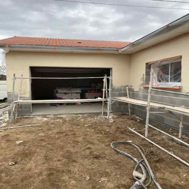Garage en construction à côté d'une maison beige, avec échafaudages, porte de garage ouverte et cour en terre battue.