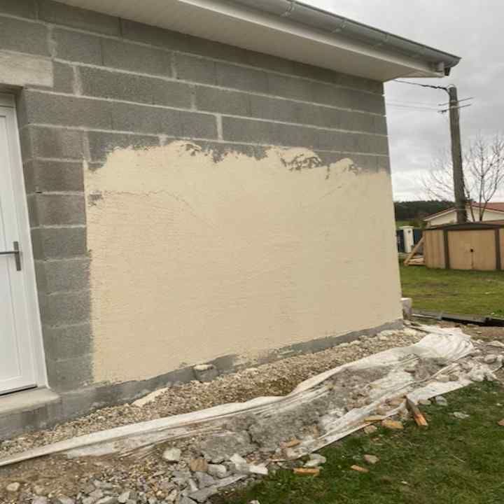 Enduit sur un mur en parpaings gris, à côté d'une porte et d'une fondation en gravier.