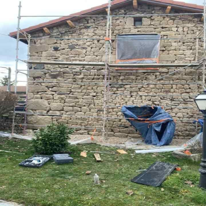 Extérieur d'une maison en pierre avec échafaudages, une petite fenêtre et des matériaux de construction dans la cour.