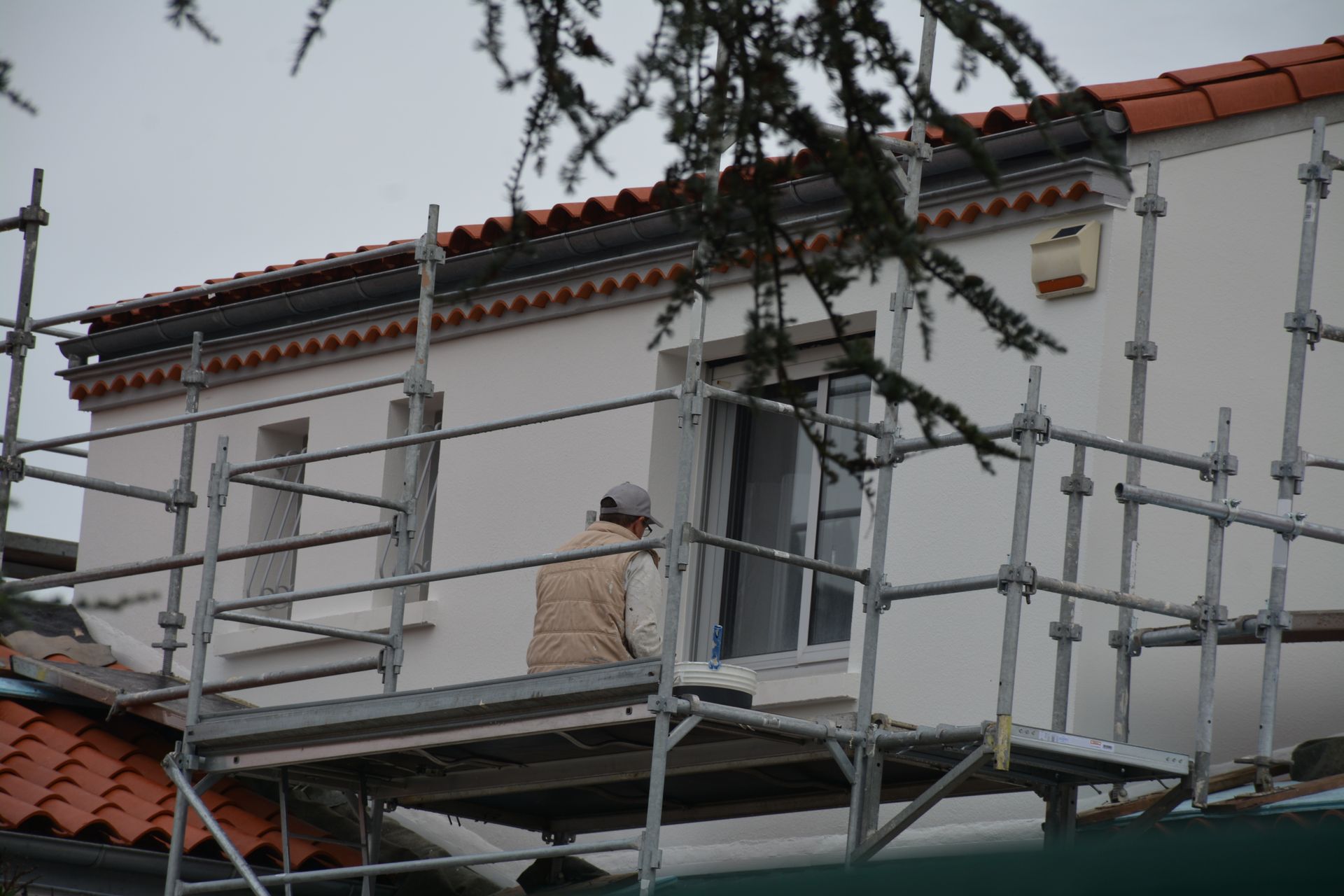 Personne sur un échafaudage près d'un bâtiment, probablement en train de peindre ou de faire des réparations. Journée nuageuse.