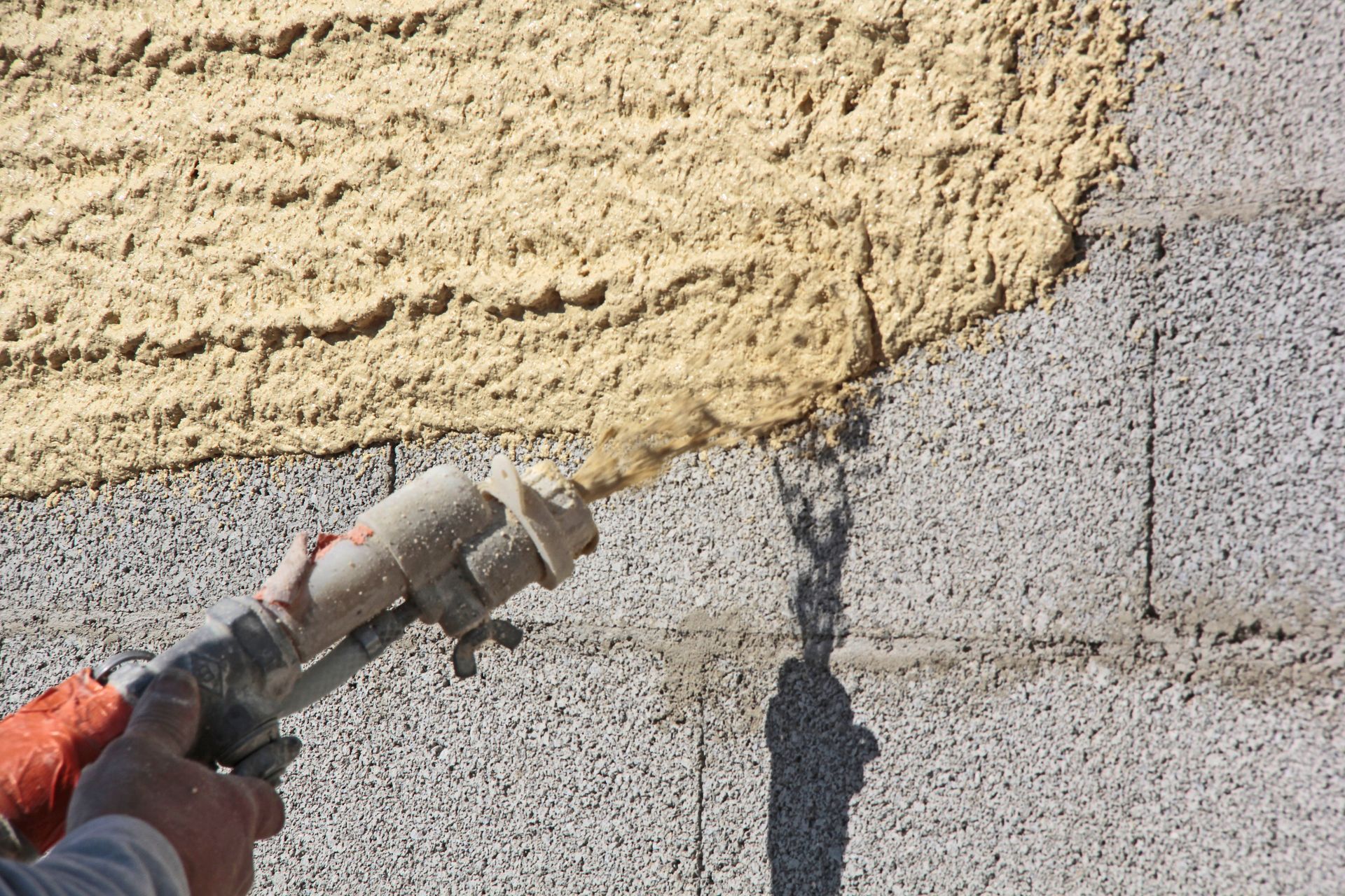 Une personne pulvérise du stuc sur un mur en parpaings ; le stuc est beige et le mur est gris.