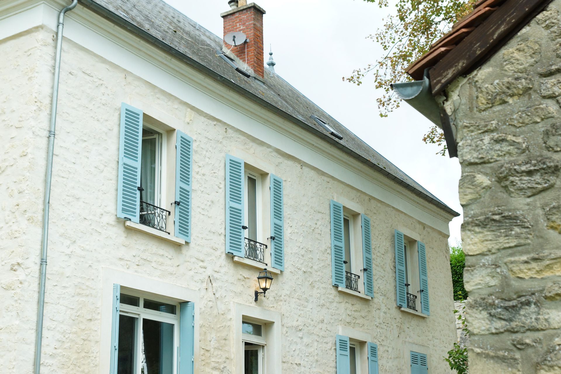 Bâtiment blanc aux volets bleu clair ; un bâtiment en pierre à droite.