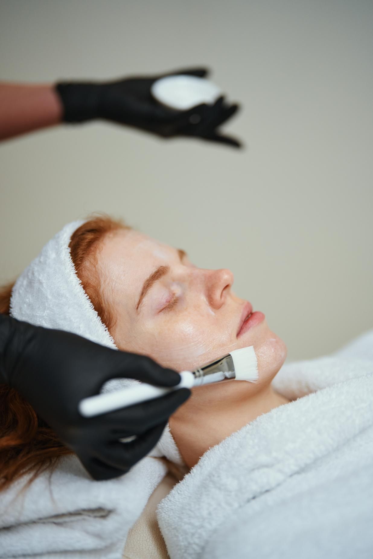 Mujer recibiendo tratamiento facial; esteticista aplicando crema con un cepillo.