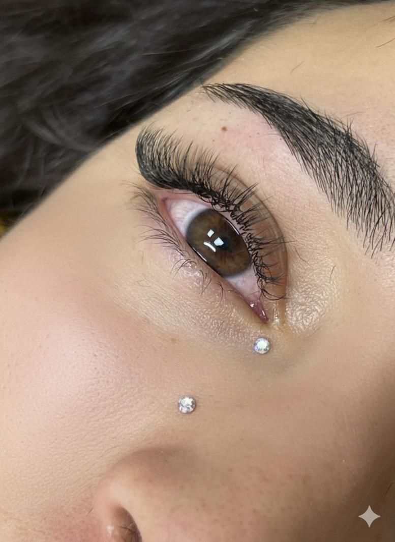 Primer plano del ojo de una persona con piercings de diamantes debajo del ojo. Tiene pestañas gruesas y cejas pobladas.