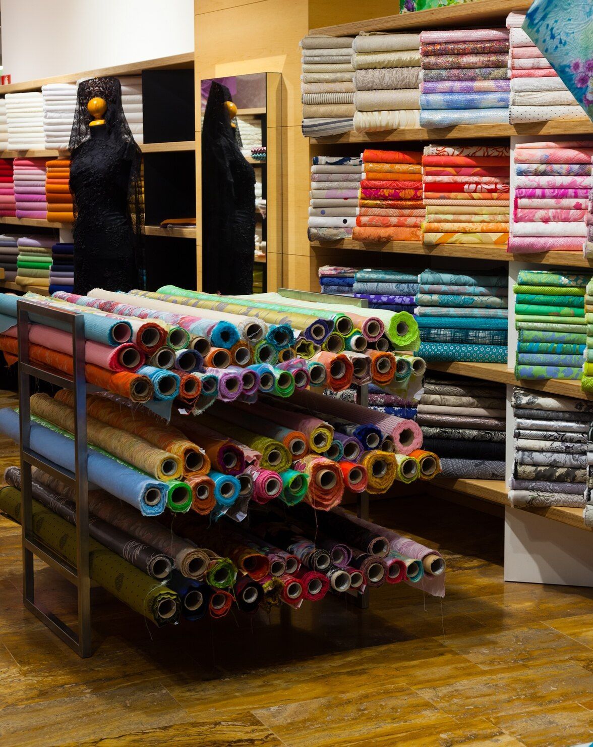 Tienda de telas con rollos de telas de colores apilados en estantes y un carrito en primer plano.