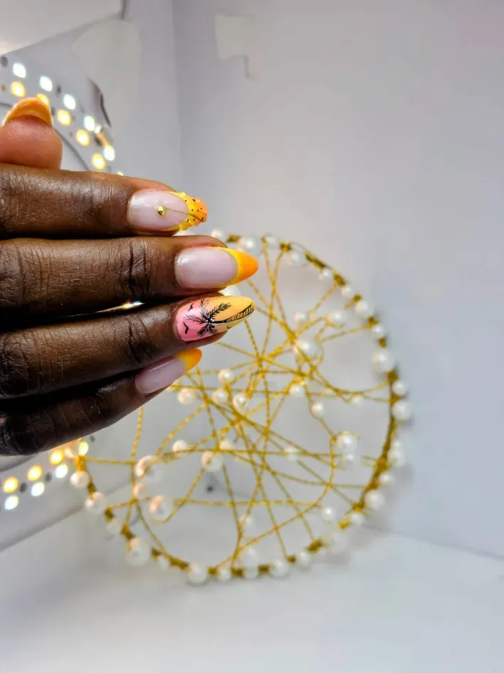 Mano con uñas decoradas en tonos nude y dorados, sostenida cerca de una decoración geométrica dorada en la pared con luces cálidas.