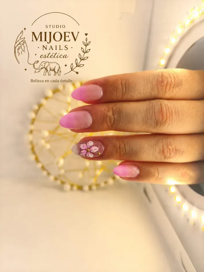 Mano con uñas decoradas con motivos florales rosas y detalles de purpurina sobre un fondo decorativo cálido.