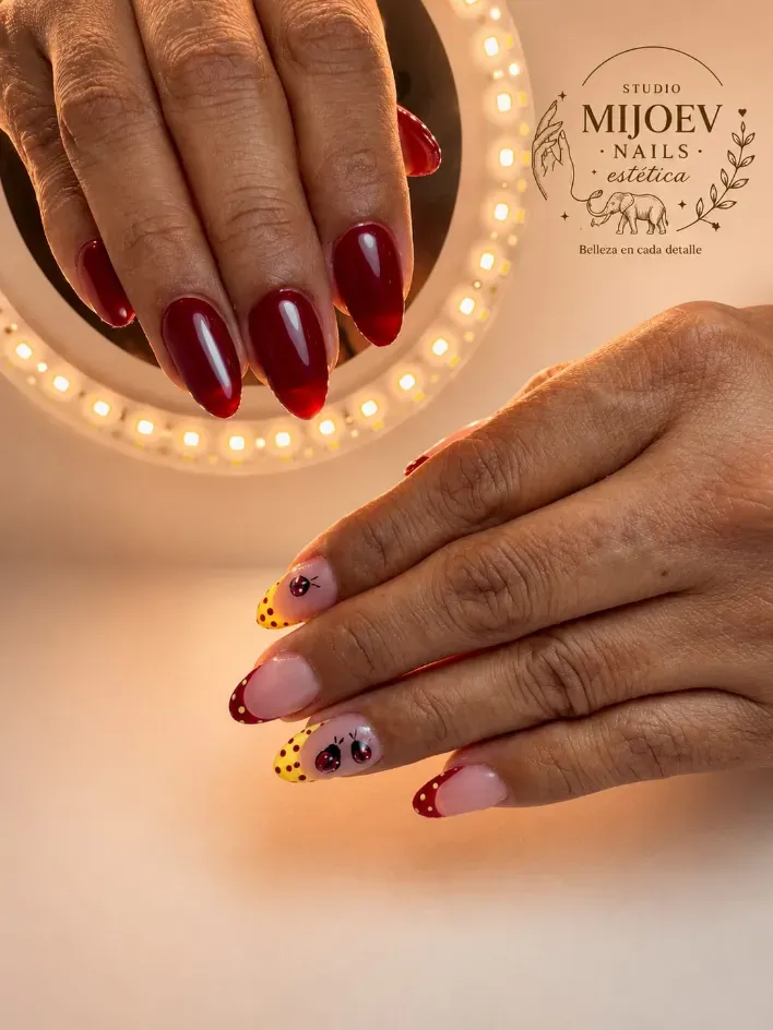 Manos que muestran uñas manicuradas en tonos burdeos y rosa con detalles dorados junto a un espejo de tocador iluminado.