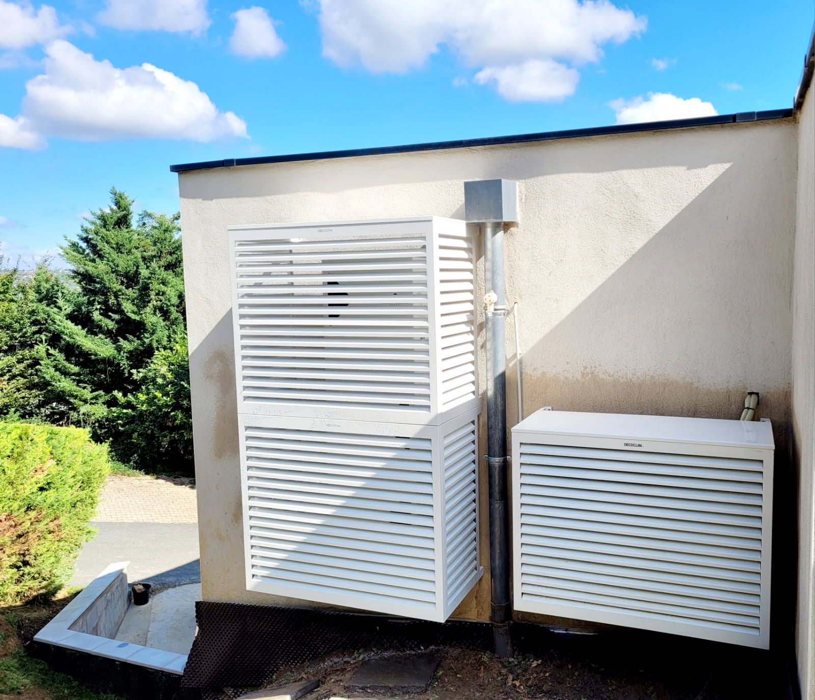 Écrans à persiennes blancs encadrant les unités extérieures sur le mur d'un bâtiment. Ciel bleu et arbres verts en arrière-plan.