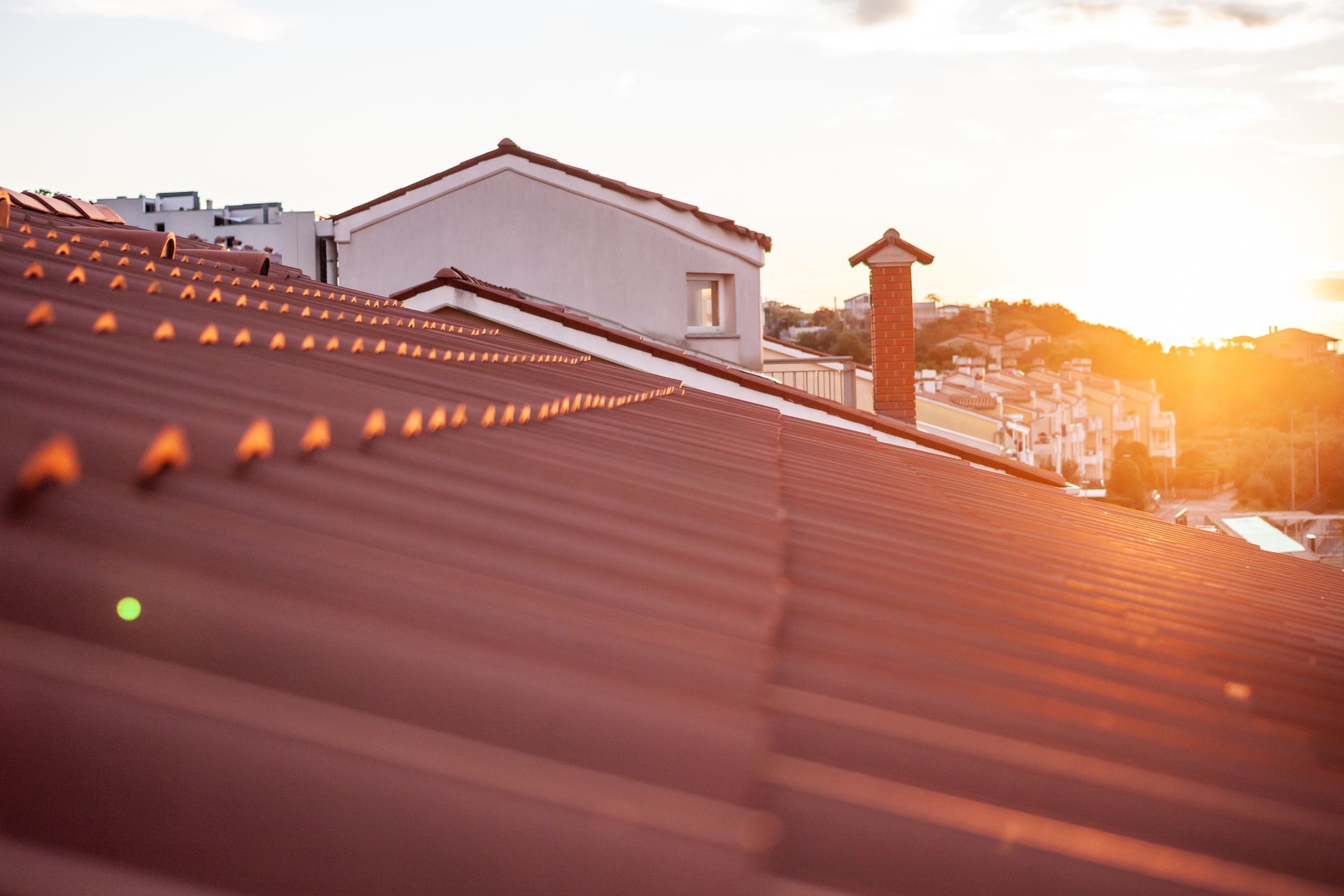 Toit de tuiles brunes avec soleil couchant en arrière-plan. Bâtiment blanc et cheminée visibles.