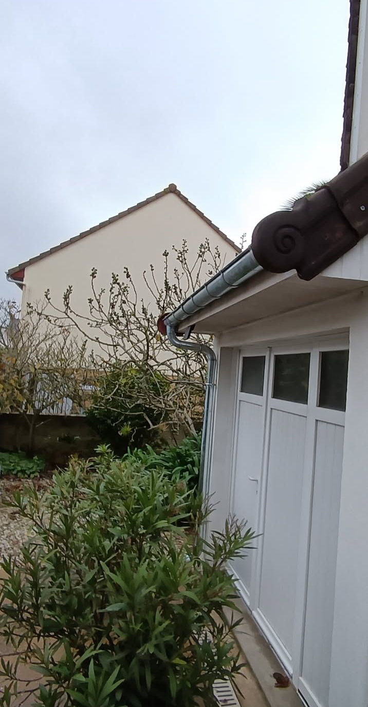Bâtiment blanc avec toit gris, gouttière et végétation. Bâtiment beige en arrière-plan, sur fond de ciel nuageux.